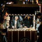 Night scene of a street vendor selling roasted chestnuts from a cart in a crowded market.