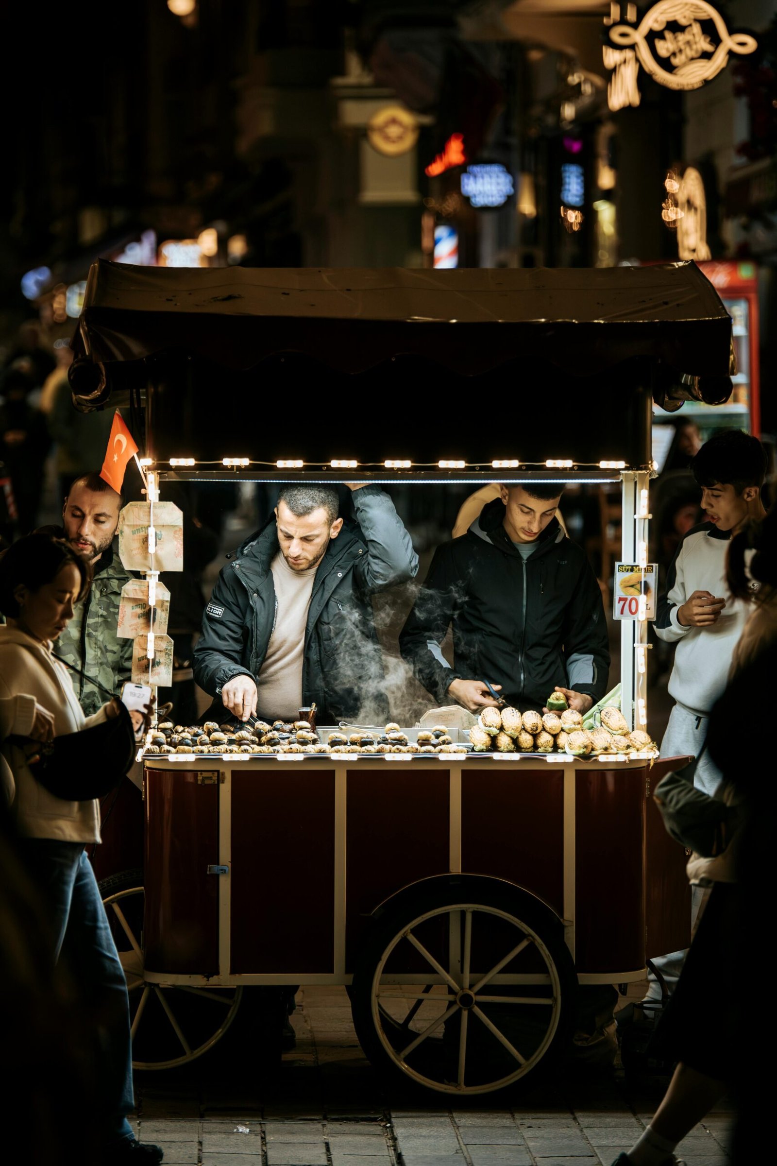 Night scene of a street vendor selling roasted chestnuts from a cart in a crowded market.